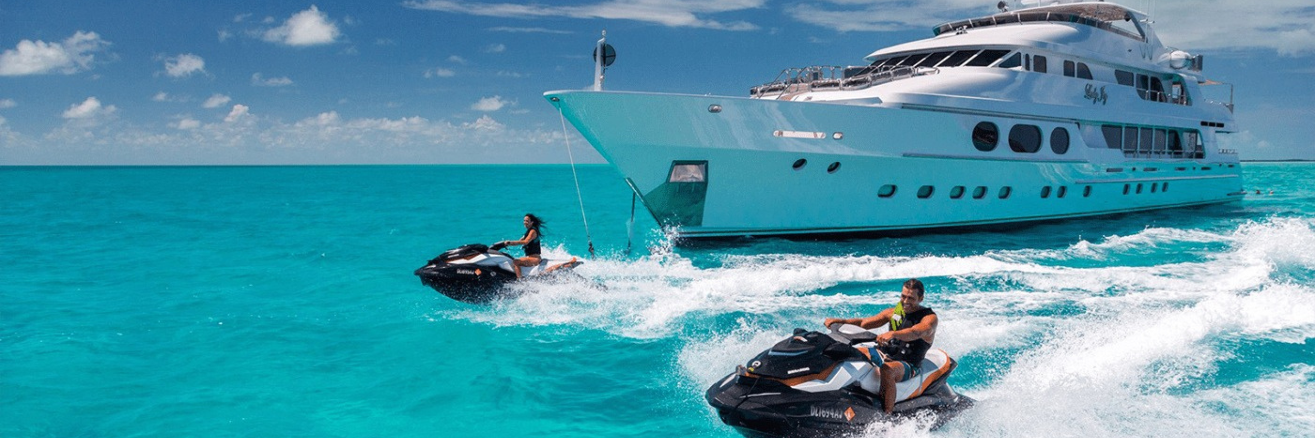 A luxury yacht anchored in clear blue waters with two jet skis speeding nearby.